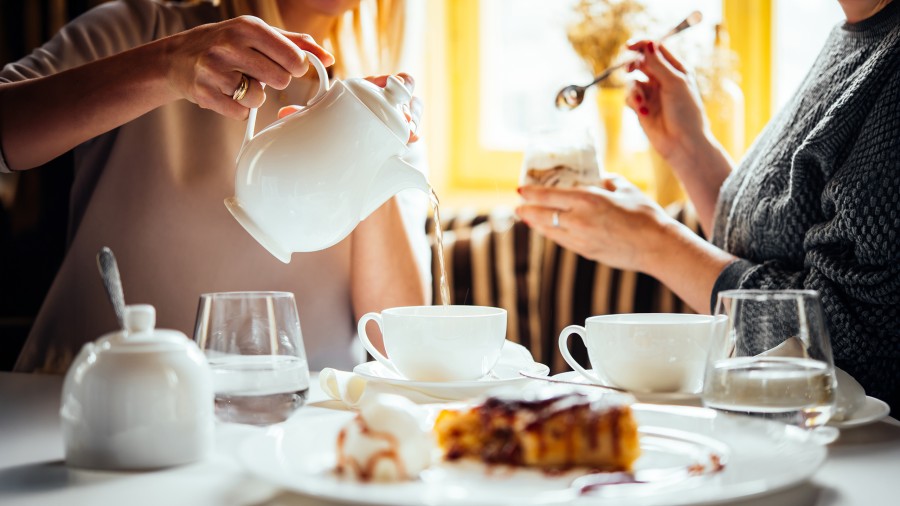 Ein mit Geschirr und Gebäck gedeckter Kaffeetisch mit zwei Personen, die man nur bis zum Halb erkennt. Eine gießt etwas aus einer Kanne in eine Tasse, die andere hält ein gefülltes Glas und einen Löffel in der Hand.